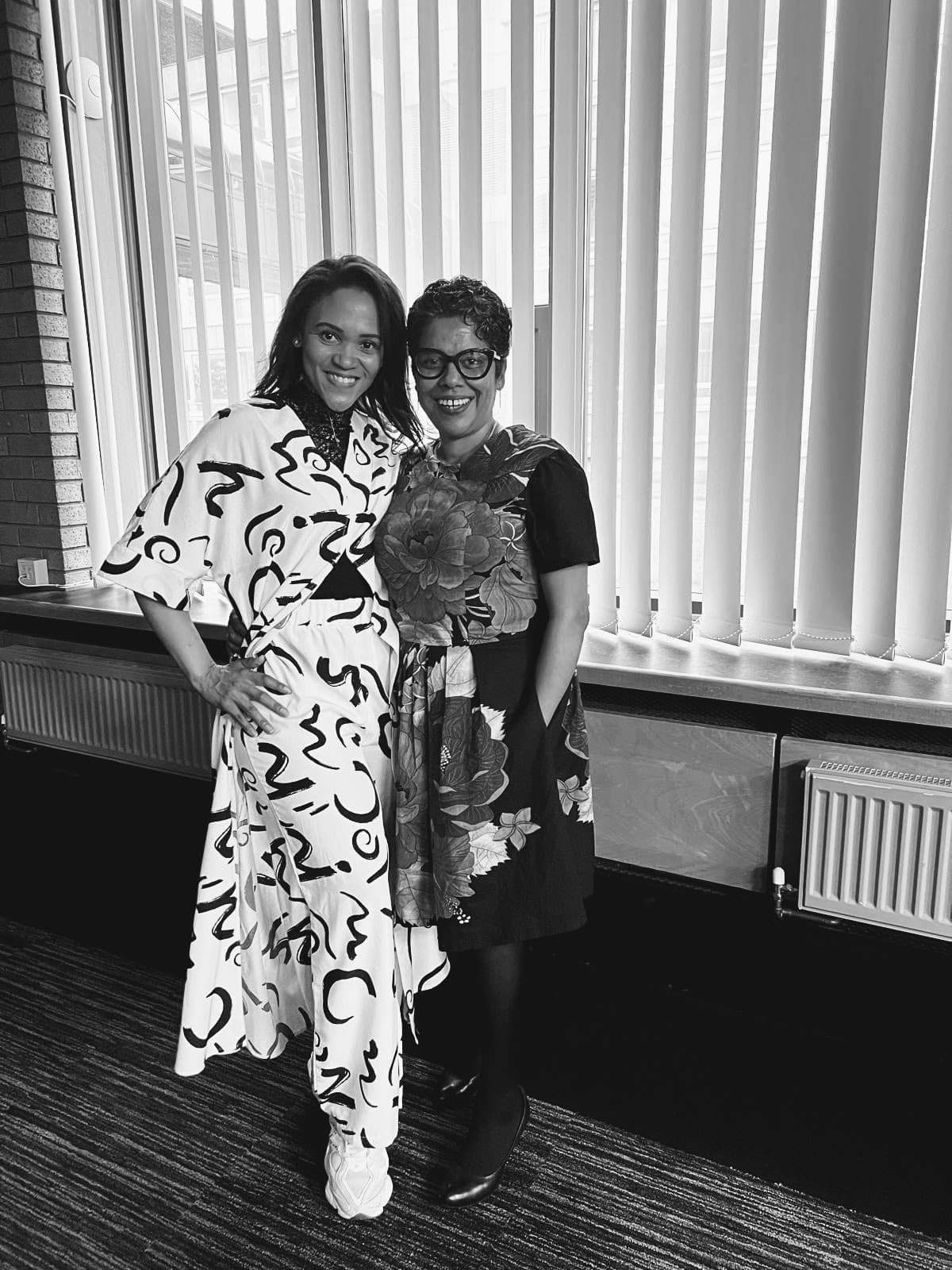 Two smiling women pose together in patterned dresses against a backdrop of vertical window blinds.
