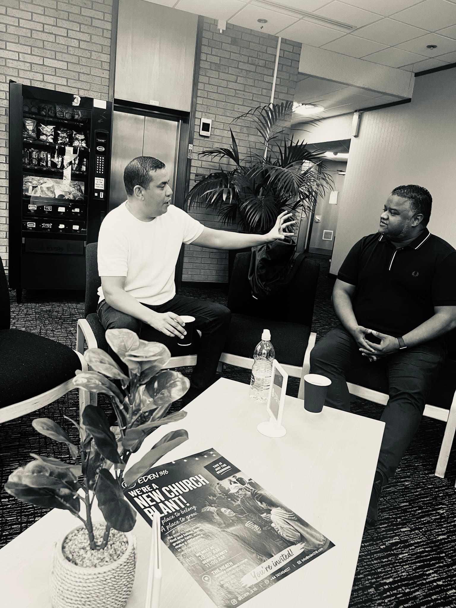 Two men conversing in a lounge area with a church flyer on the table.