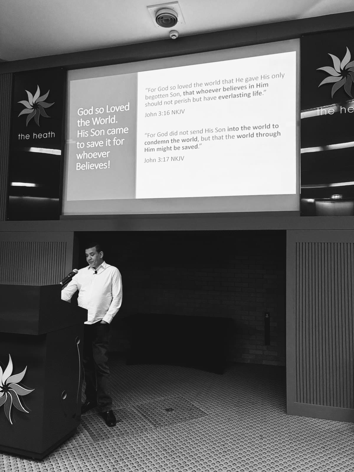 Man at a podium speaking before a screen displaying Bible verses from John 3:16-17.
