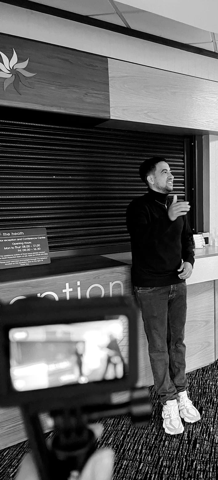 Man in a turtleneck gestures while being filmed in front of a reception desk.