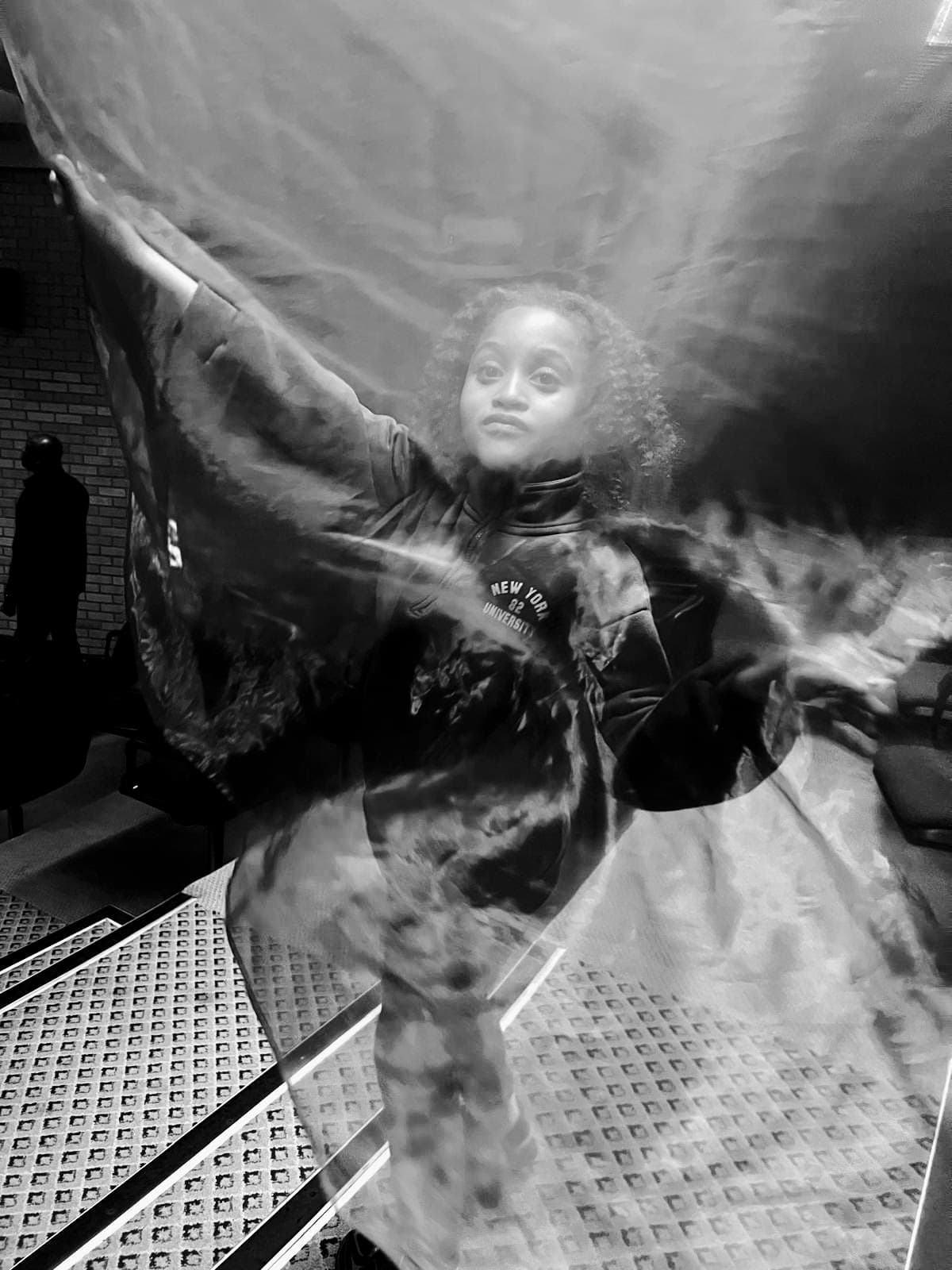 Person in NYU sweatshirt behind flowing, translucent fabric with arms raised, in black and white.