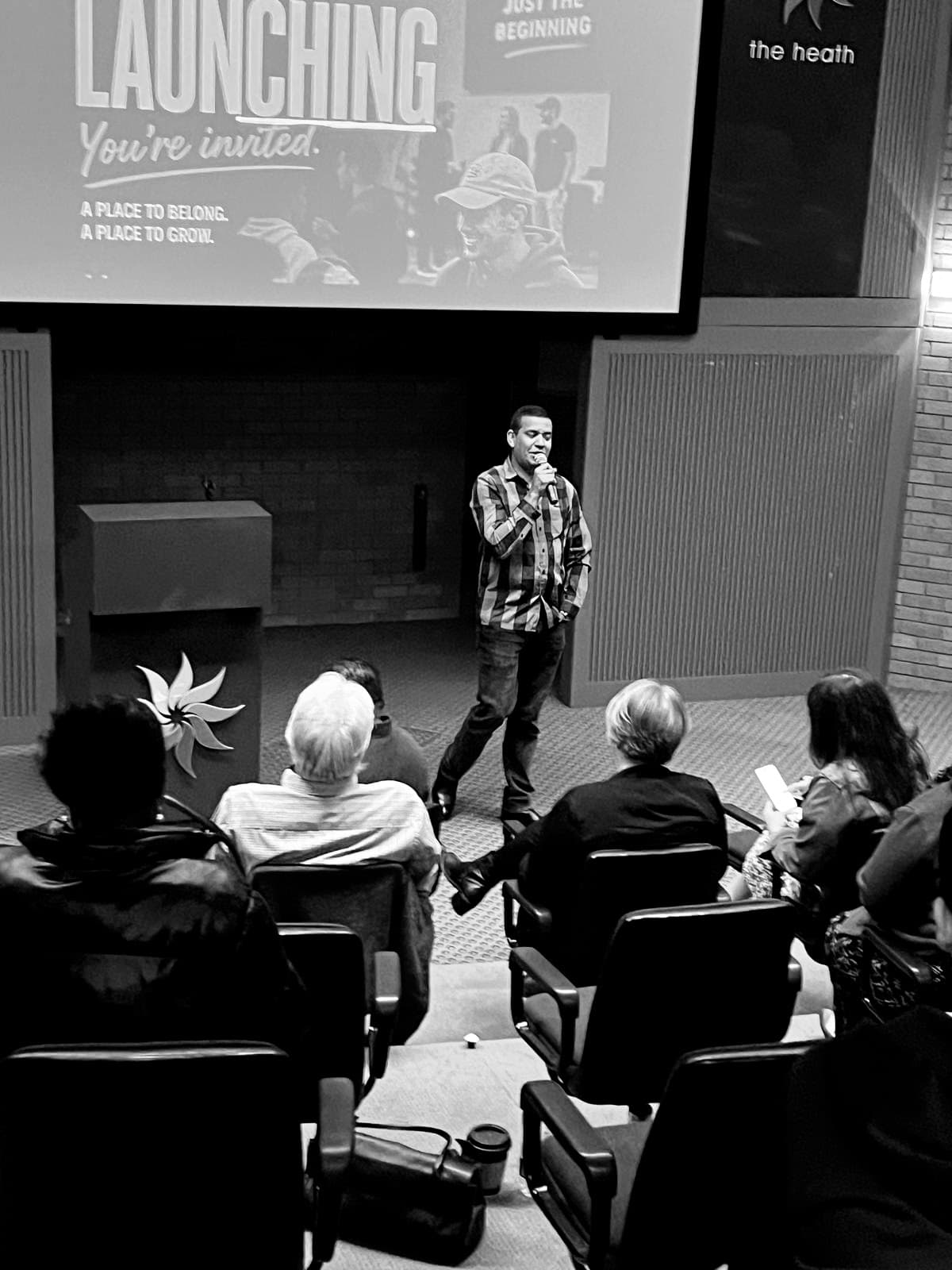 Man in plaid shirt speaking into a microphone on stage before a seated audience.