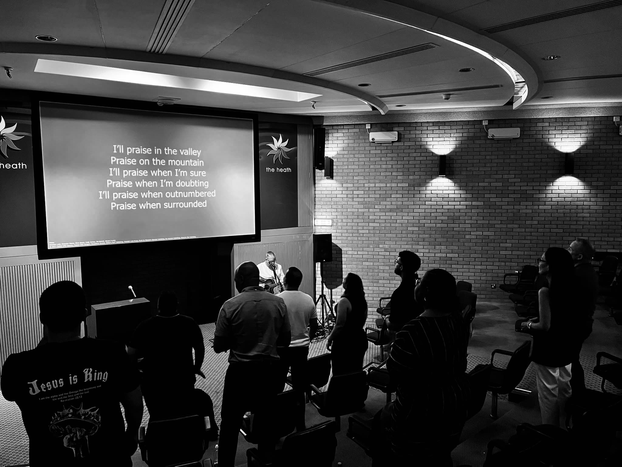 People stand in an auditorium singing worship lyrics displayed on a large screen.