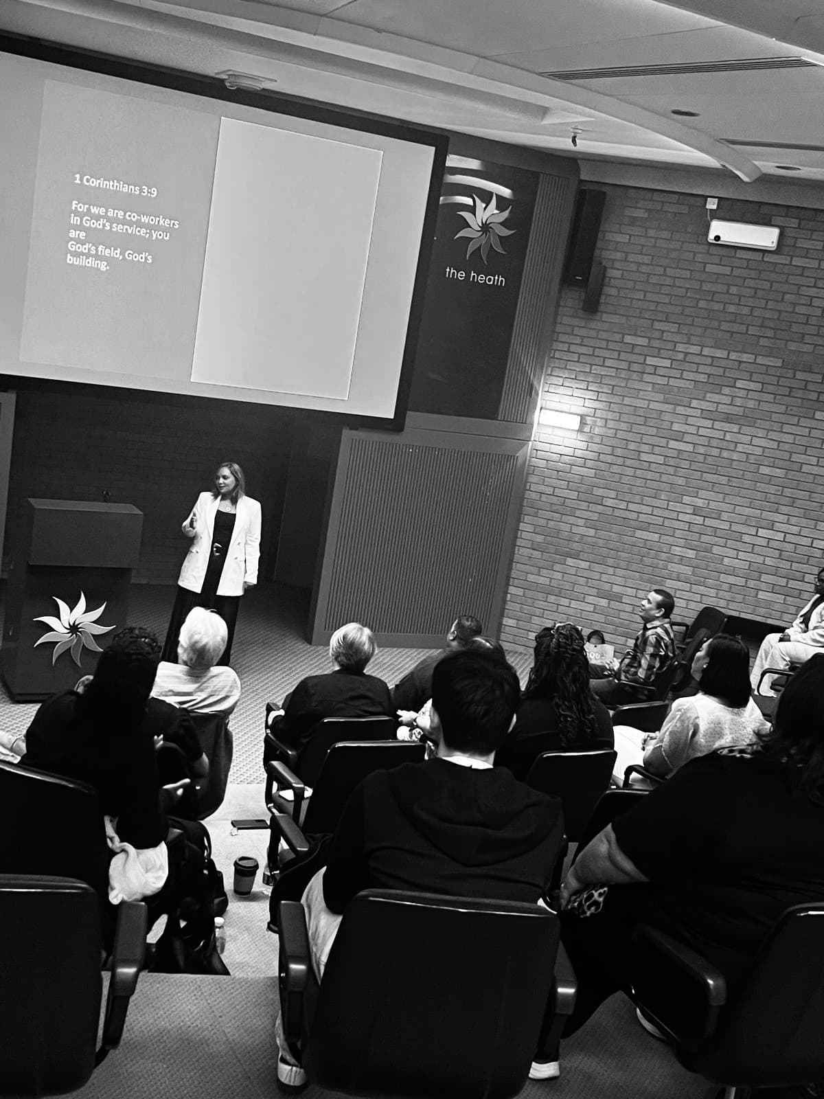 Woman in white jacket presenting a Bible verse to a seated audience in an auditorium.