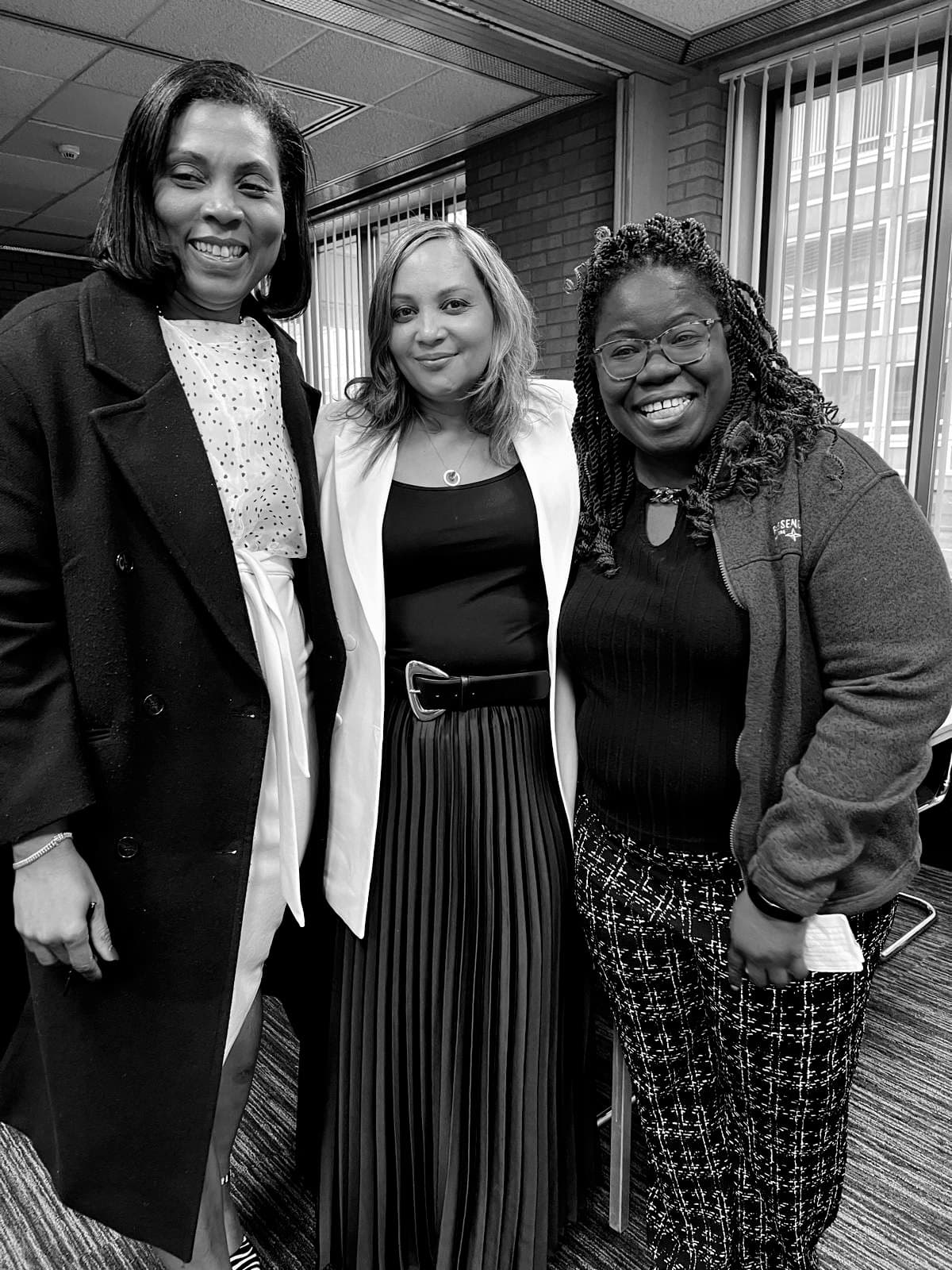 Three smiling women in professional attire pose together in a black and white indoor setting.