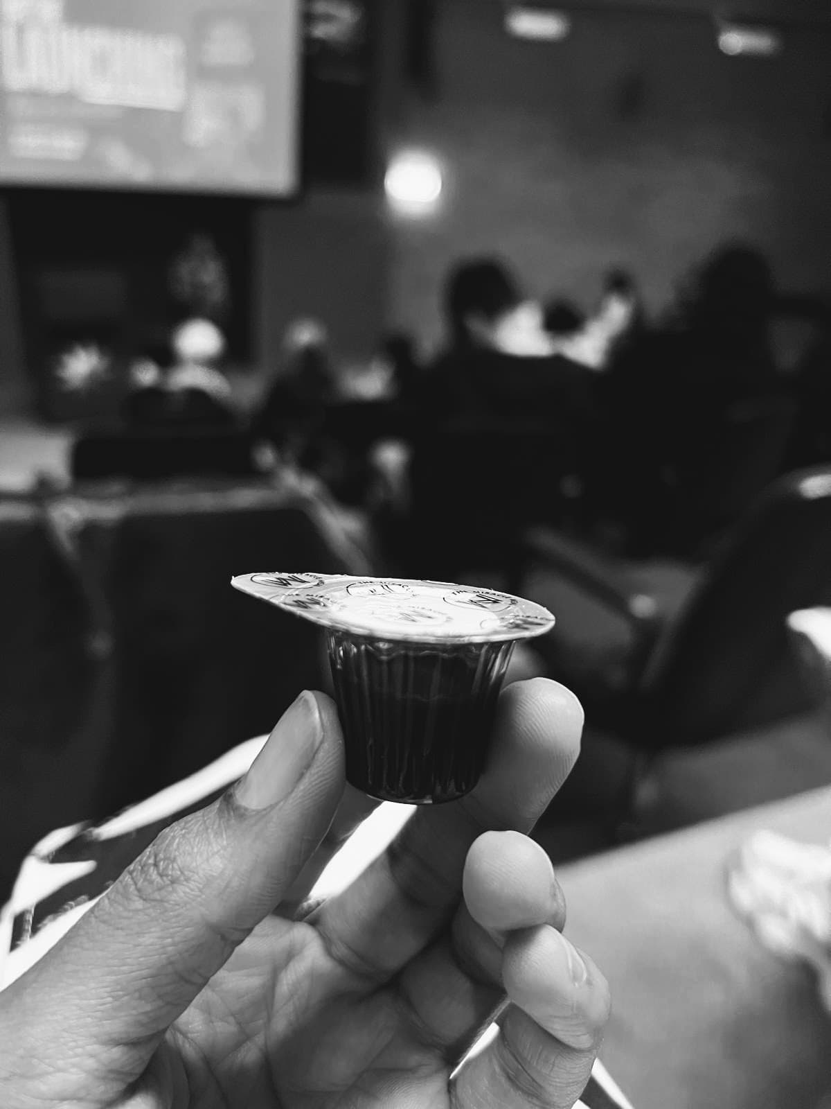 Hand holding a small communion cup in a black and white, blurred auditorium setting.